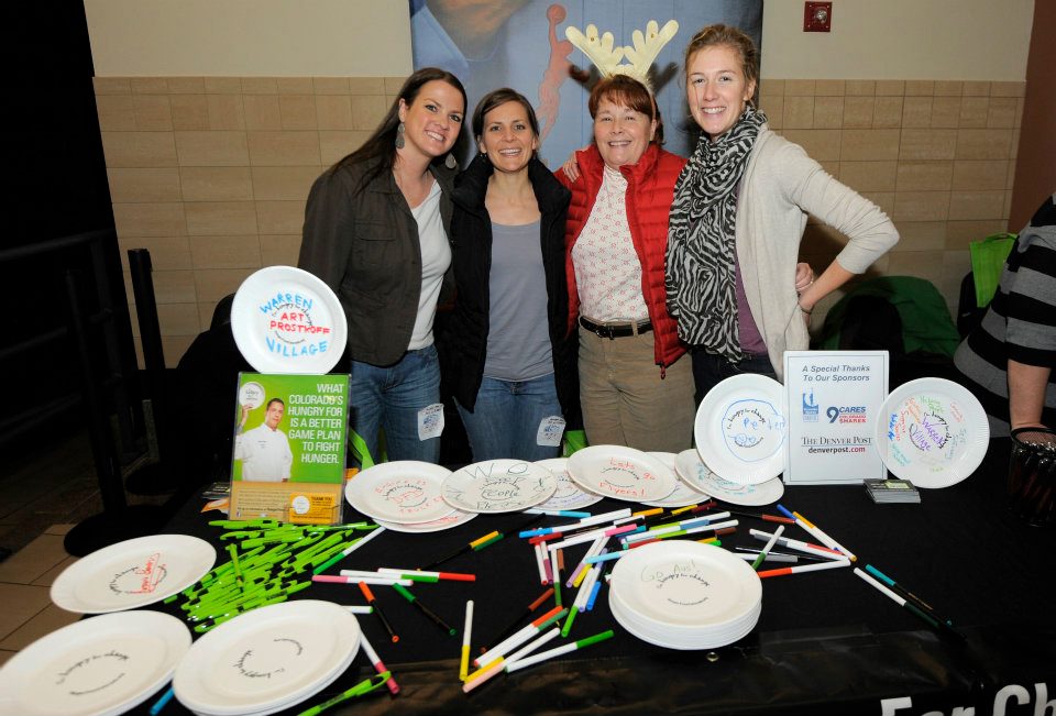 Four women smile and pose for a portrait (one is wearing a red puffer vest and reindeer antlers) in front of a table with paper plates and markers. Some of the plates have drawings on them and all read, "I'm hungry for change."