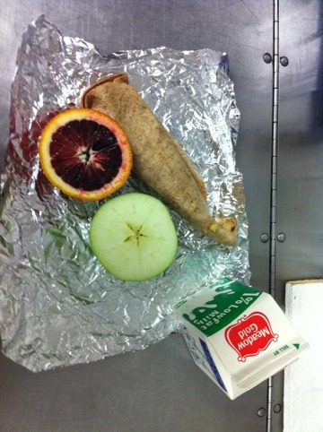 Lunch consisting of a green apple and blood orange (sliced in half), and a wrap on unwrapped tin foil and a carton of 1% low fat milk