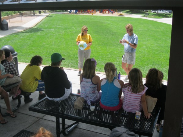 Two adults (left holds a globe, right holds a piece of paper) present to students siting on picnic tables in an outdoor shelter near a playground