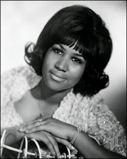 black and white portrait of Aretha Franklin leaning against the back of a wire chair in a sparkly dress