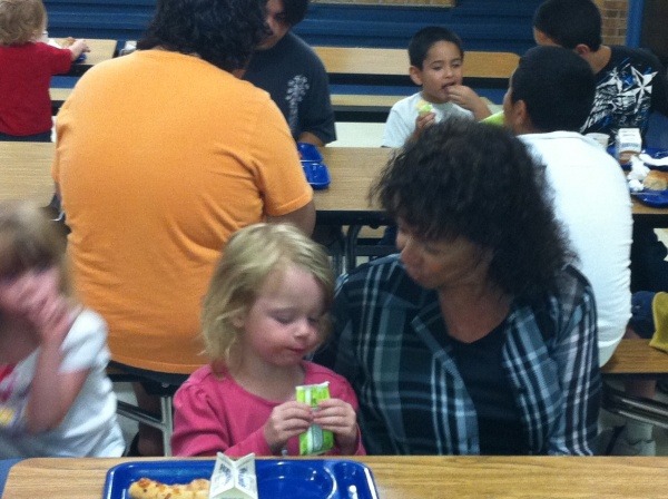 A woman in a black t-shirt and blue plaid flannel speaks to a child in pink who is eating her lunch