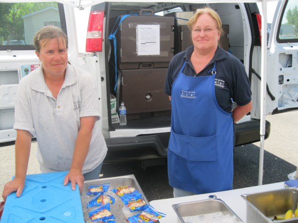 Two women (left is wearing gray polo, right is wearing a blue polo and blue apron) standing behind a covered table serving food.