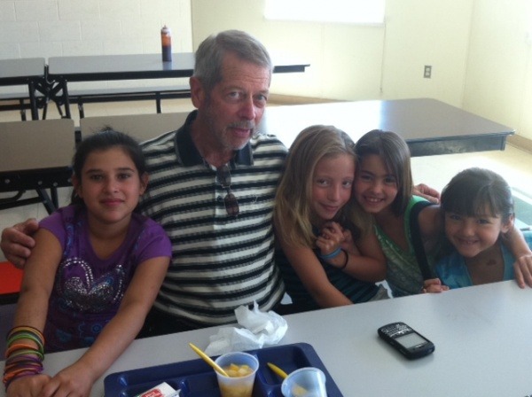 Man in striped blue and white shirt poses with 4 young girls (some smiling) at a school lunch table