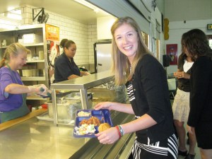 woman poses with a lunch tray, background is others waiting in line to be served and two lunch ladies in a school cafeteria
