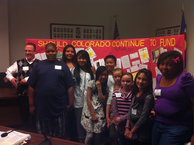 Students an a teacher standing in front of a red poster that reads "Should Colorado Continue to fund our policy"