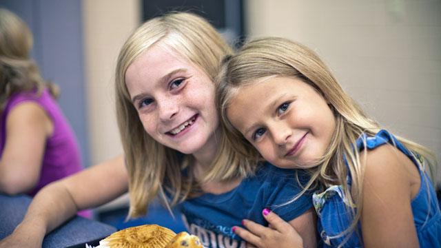 Two young girls (left is wearing a blue shirt and right is wearing a blue dress) smiling. The girl on the right rests her head on the shoulder of her friend. Both are smiling.