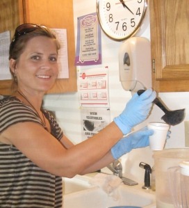 woman in stripped shirt and blue medical gloves ladles liquid into a small Styrofoam cup