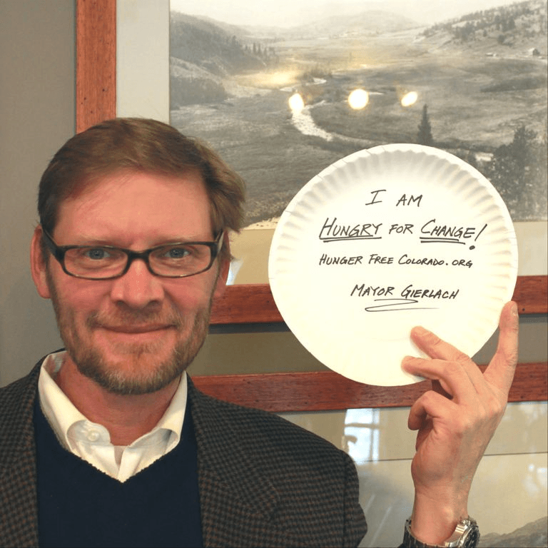 Man in blazer holds plate that says "I am HUNGRY FOR CHANGE! Hunger Free Colorado.org Mayor Gierlach"